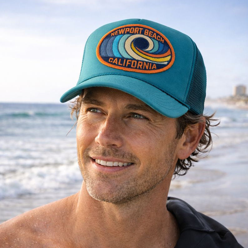 Man wearing a Port Sandz jade colored trucker hat with 'Newport Beach, California' embroidered on the patch on the front of the hat. He's got a wetsuit over his shoulder and a surfboard at the beach.