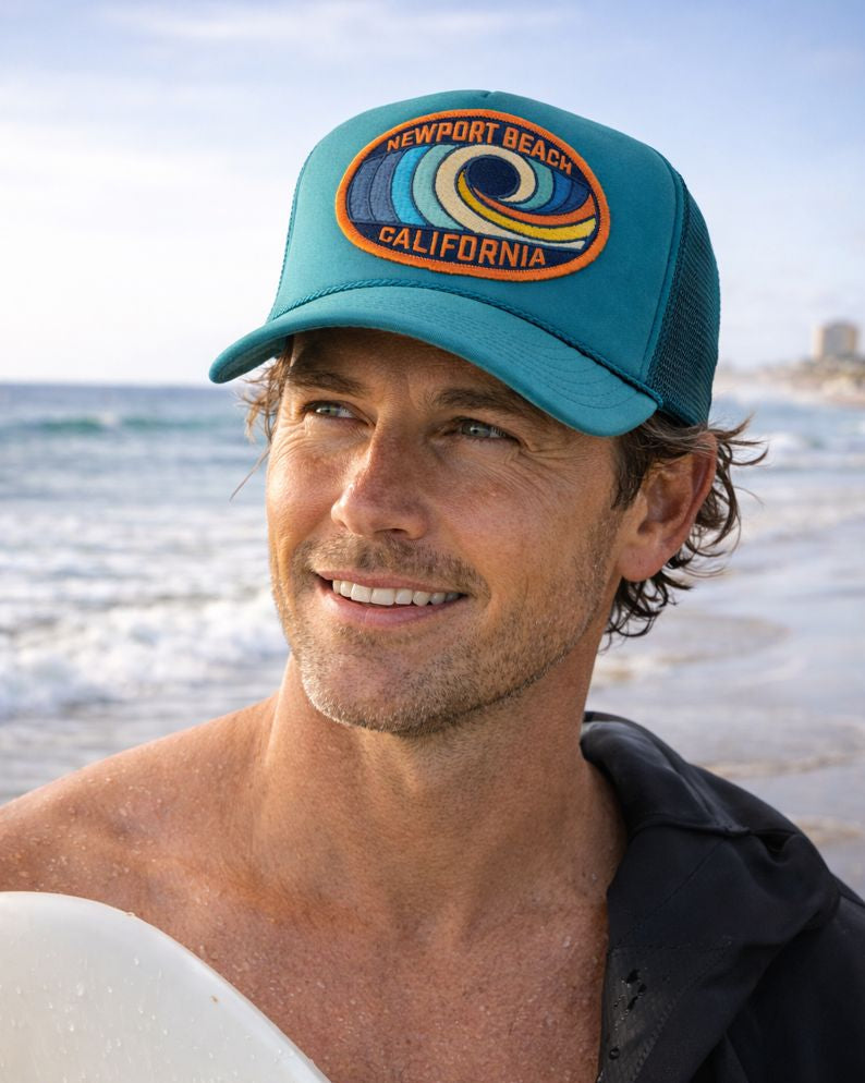 Man wearing a Port Sandz jade colored trucker hat with 'Newport Beach, California' embroidered on the patch on the front of the hat. He's got a wetsuit over his shoulder  and a surfboard at the beach.