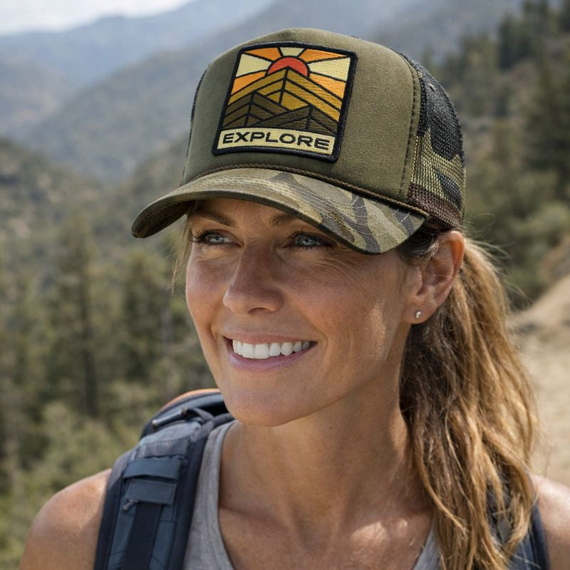 Woman wearing a Port Sandz trucker hat with 'EXPLORE' embroidered on the front panel patch in a mountainous outdoor setting