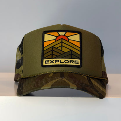 Camo colored trucker hat with viscose embroidered patch that reads "EXPLORE" on the front panel. 