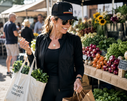 Woman shopping at a farmers market with produce and reusable bags wearing Port Sandz trucker hat that reads: I'm Busy