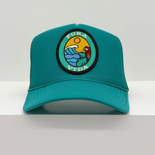 Port Sandz trucker hat in jade color with viscose embroidered oval patch reading PURA VIDA and image of colorful bird in tree overlooking ocean waves and a sun in the background.