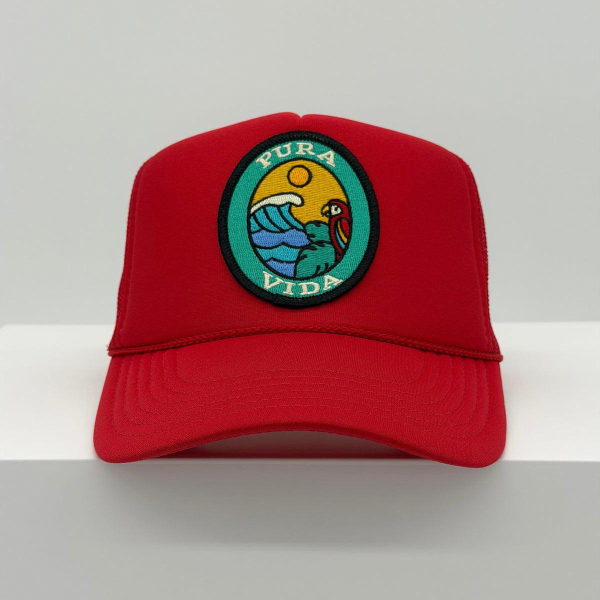 Port Sandz trucker hat in red color with viscose embroidered oval patch reading PURA VIDA and image of colorful bird in tree overlooking ocean waves and a sun in the background.