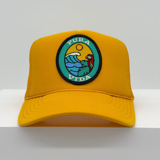 Port Sandz trucker hat in yellow color with viscose embroidered oval patch reading PURA VIDA and image of colorful bird in tree overlooking ocean waves and a sun in the background.